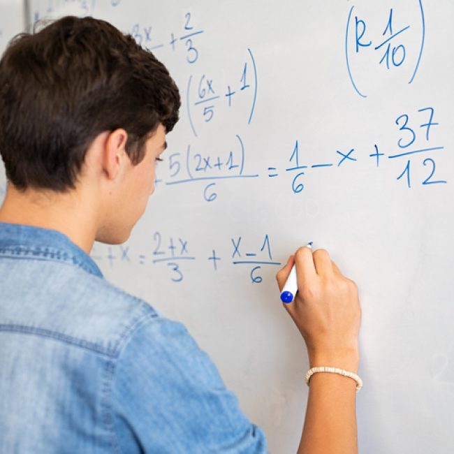Back view of high school student solving math problem on whiteboard in classroom. Young man writing math solution on white board using marker. College guy solving math expression during lesson.