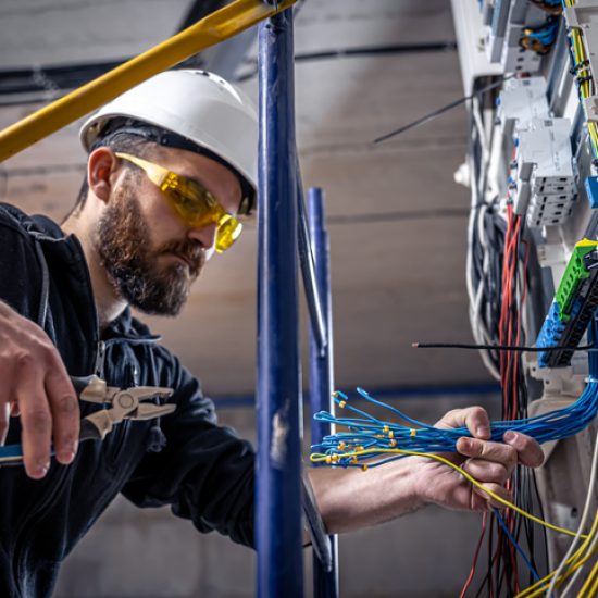 A male electrician works in a switchboard with an electrical connecting cable, connects the equipment with tools.